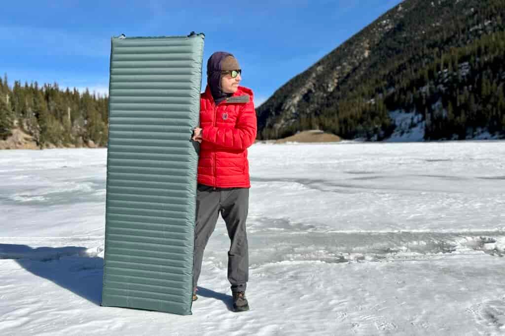 A man holds a camping mattress vertically in a snowy mountain setting.