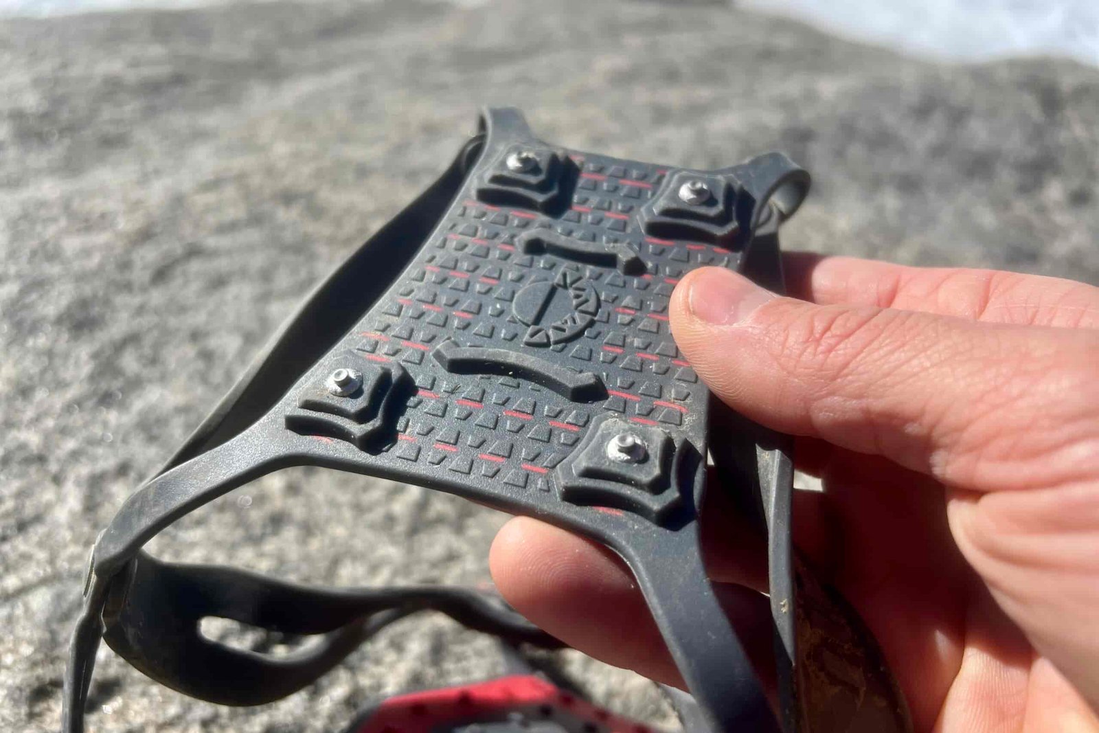 A hand shows one of the traction zones on an ice cleat with a rock background.