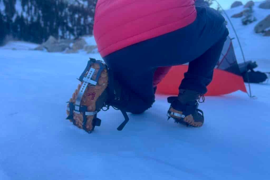 A hiker kneels in snow, showing the bottom of his foot wearing an aggressive trail crampon.