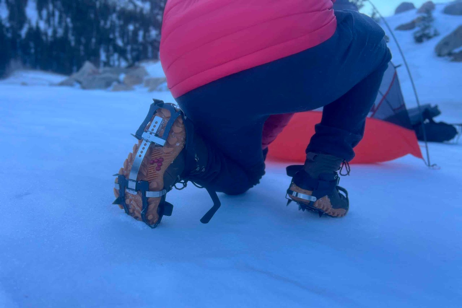 A hiker kneels in snow, showing the bottom of his foot wearing an aggressive trail crampon.