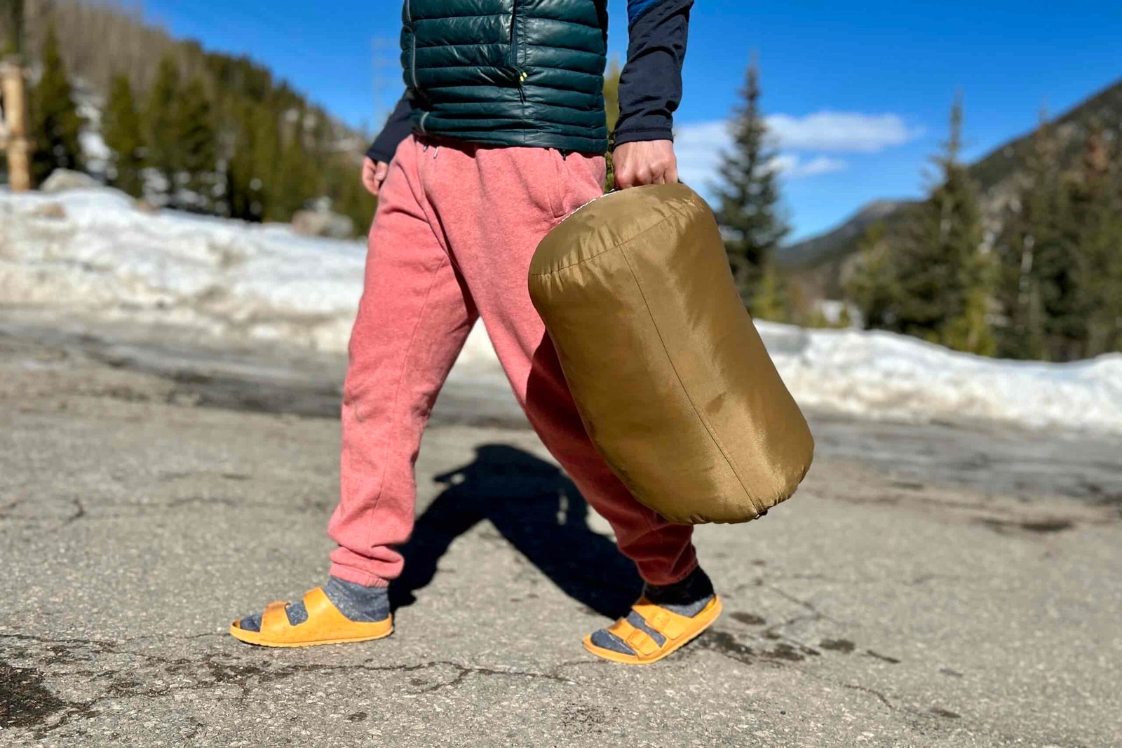 A person walks through a snowy parking lot carrying a sleeping bag in a stuff sack at their side.