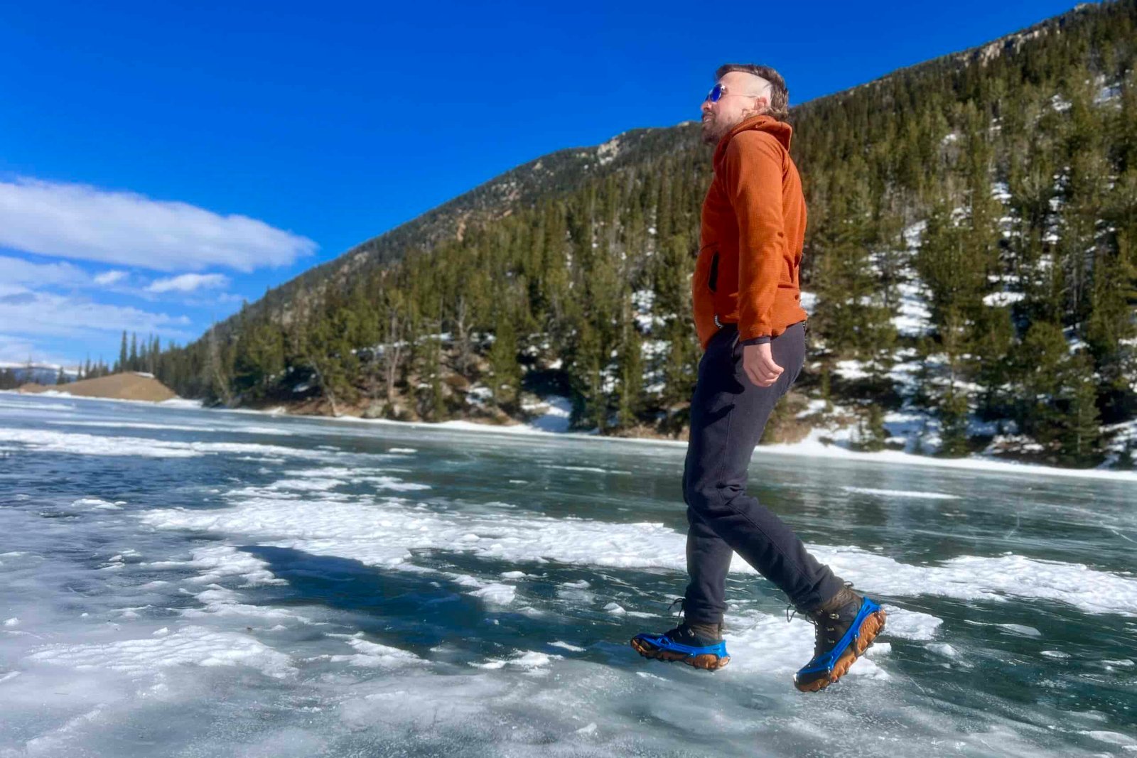 A hiker walks across an icy lake on a sunny day while wearing ice cleats. Trees in the background.