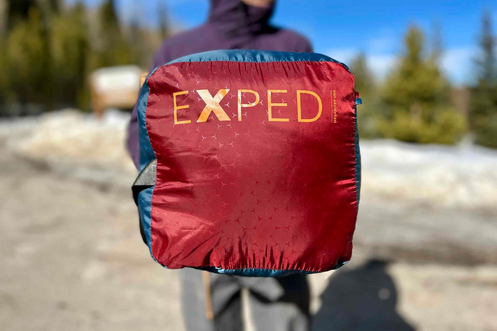 A person holds a sleeping bag in a stuff sack under their arm so the logo is visible.