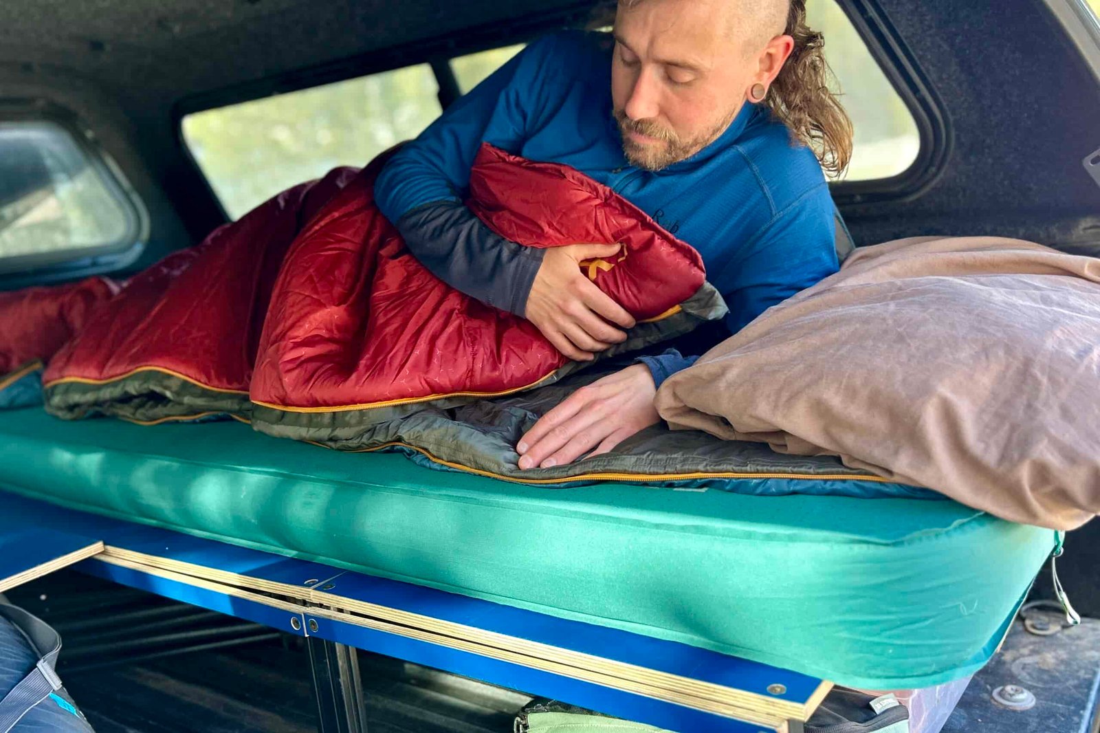 A man in a sleeping bag on a camping mattress in the back of a truck.