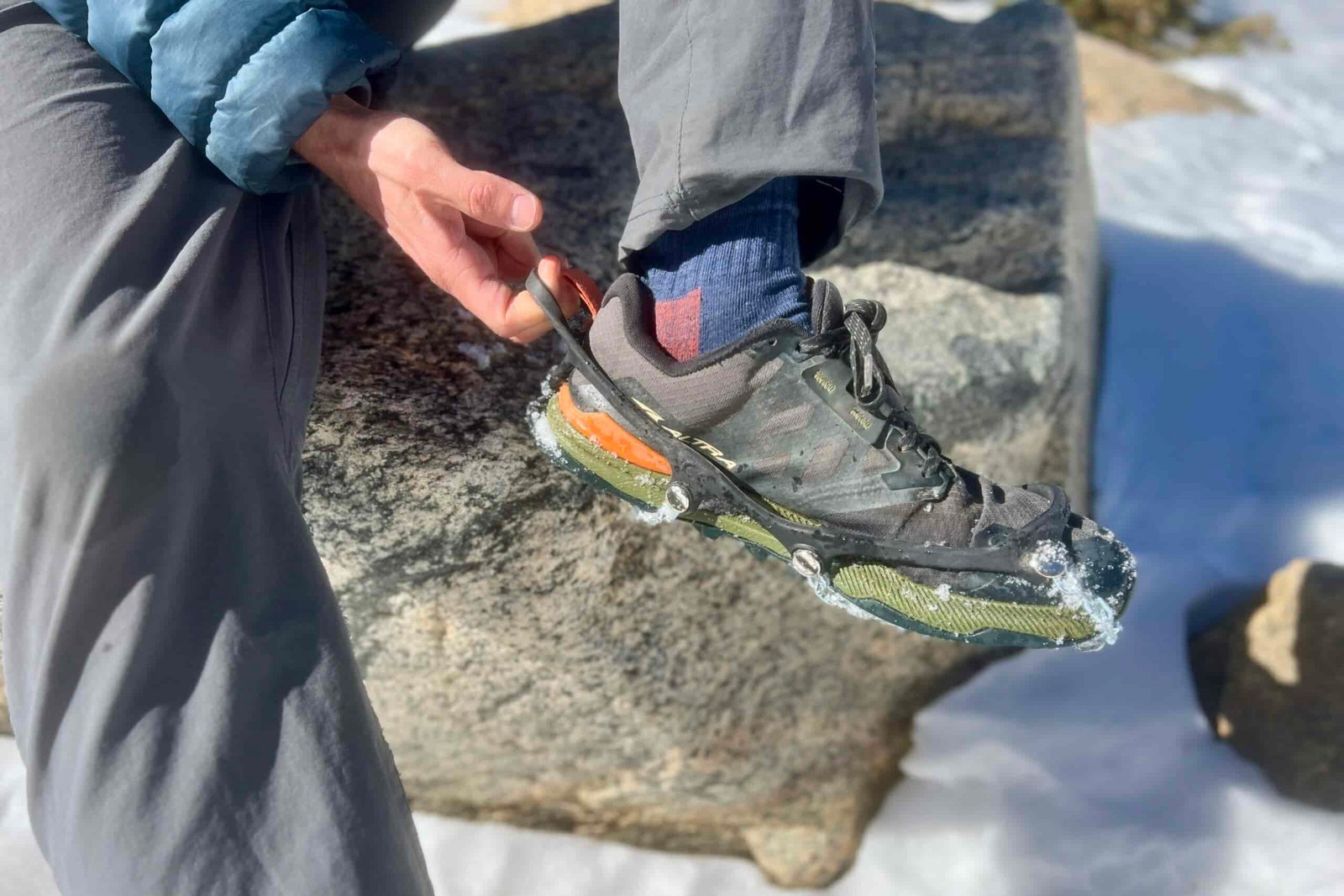 A hiker sits on a rock and pulls a pair of ice cleats onto his feet.