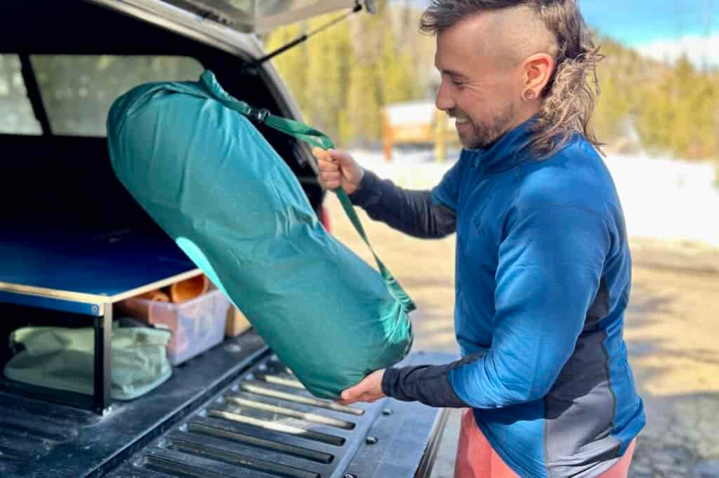 A man smiles as he sets the large storage duffel of a camping mattress in the back of a truck.