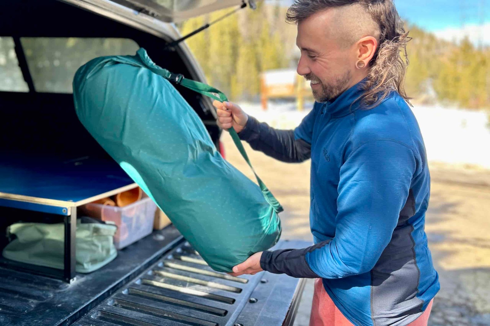 A man smiles as he sets the large storage duffel of a camping mattress in the back of a truck.