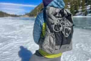 A hiker looks away from the camera on a frozen lake while wearing a backpack with ice cleats attached to the outside.