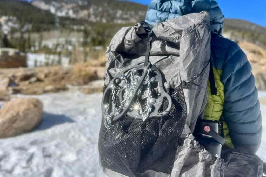 A hiker facing away from the camera wearing a backpack with a pair of ice cleats attached to the outside. Snowy forest background.