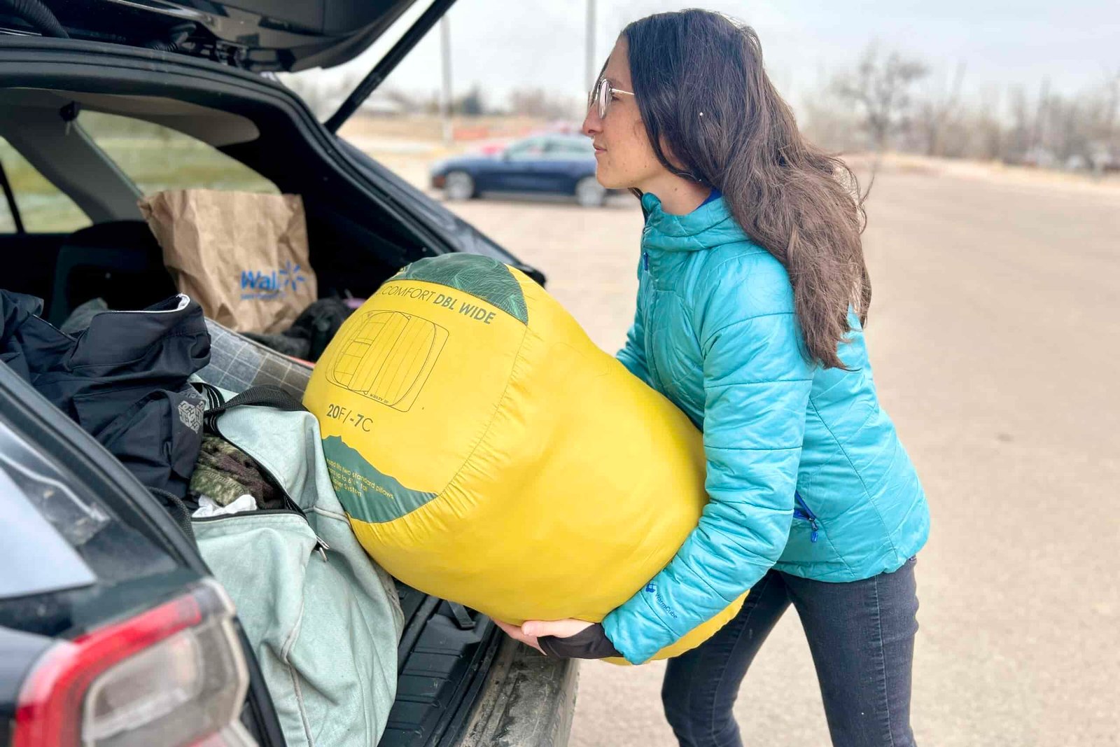 A woman sets a large sleeping bag in the back of a car with a full trunk.