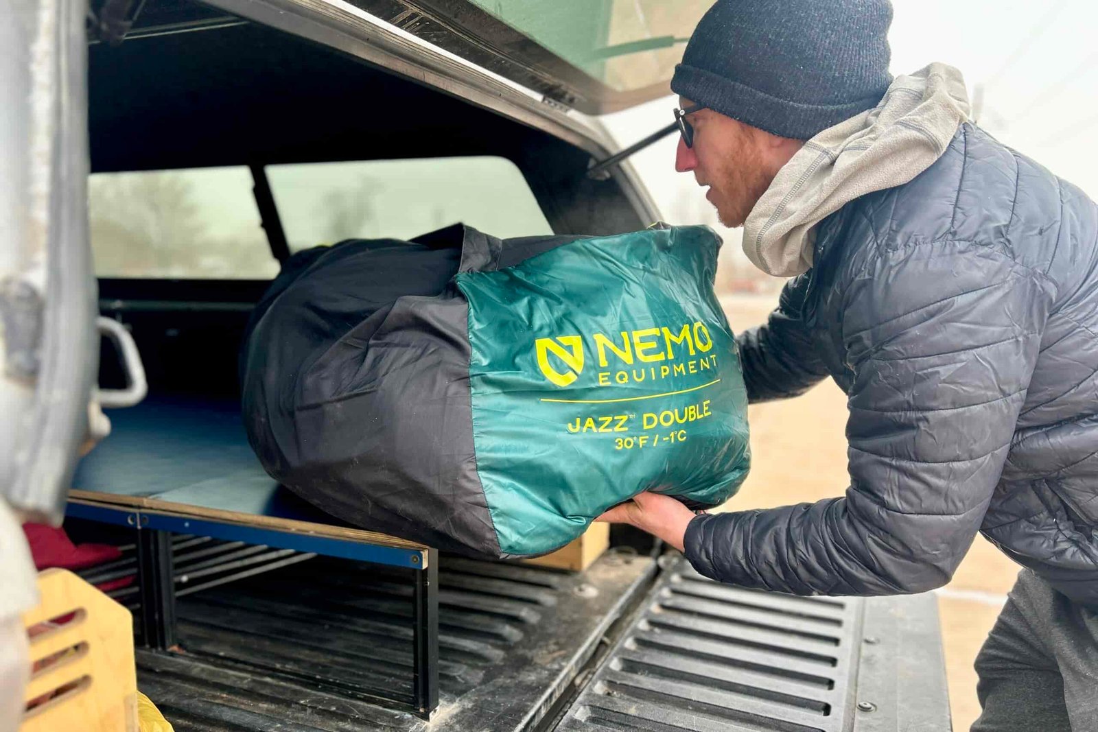 A man in a puffy jacket and hat sets a large sleeping bag in the back of a truck.