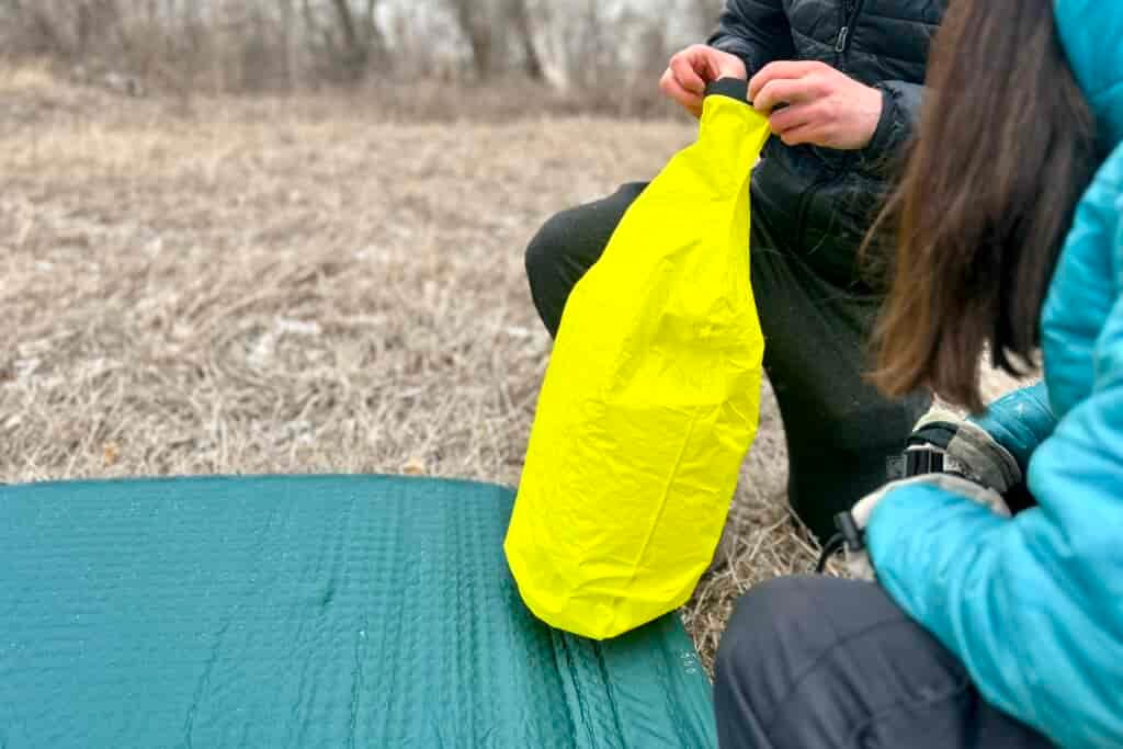 Two people kneel next to a camping mattress as one of them inflates a stuff sack attached to the side.