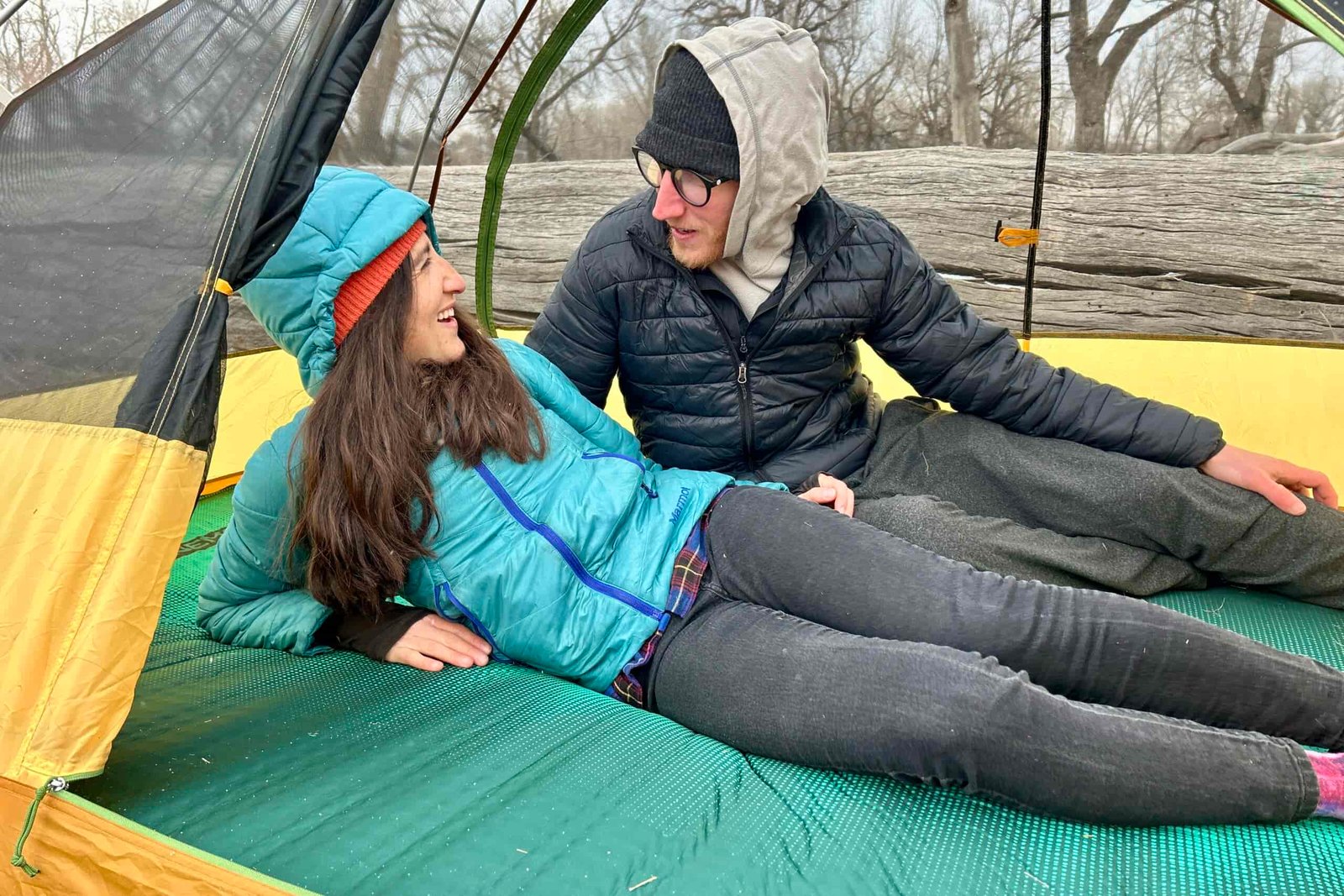 A woman and man are smiling and talking while laying on a camping mattress in a tent in the woods.