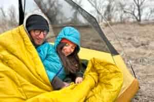 A man and woman are smiling and laying in a double sleeping bag in a tent.