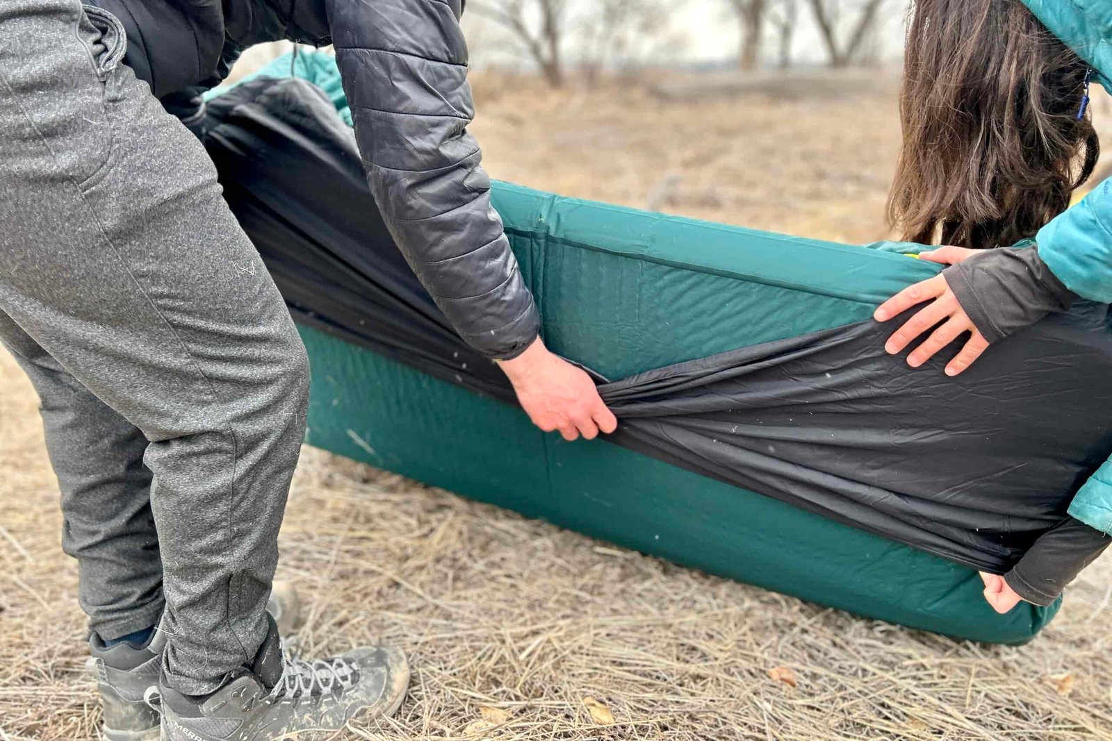 Two people insert a camping mattress into the mattress sleeve of a sleeping bag.