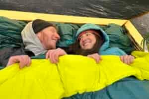 A man and woman smile at each other in a double sleeping bag in a tent.