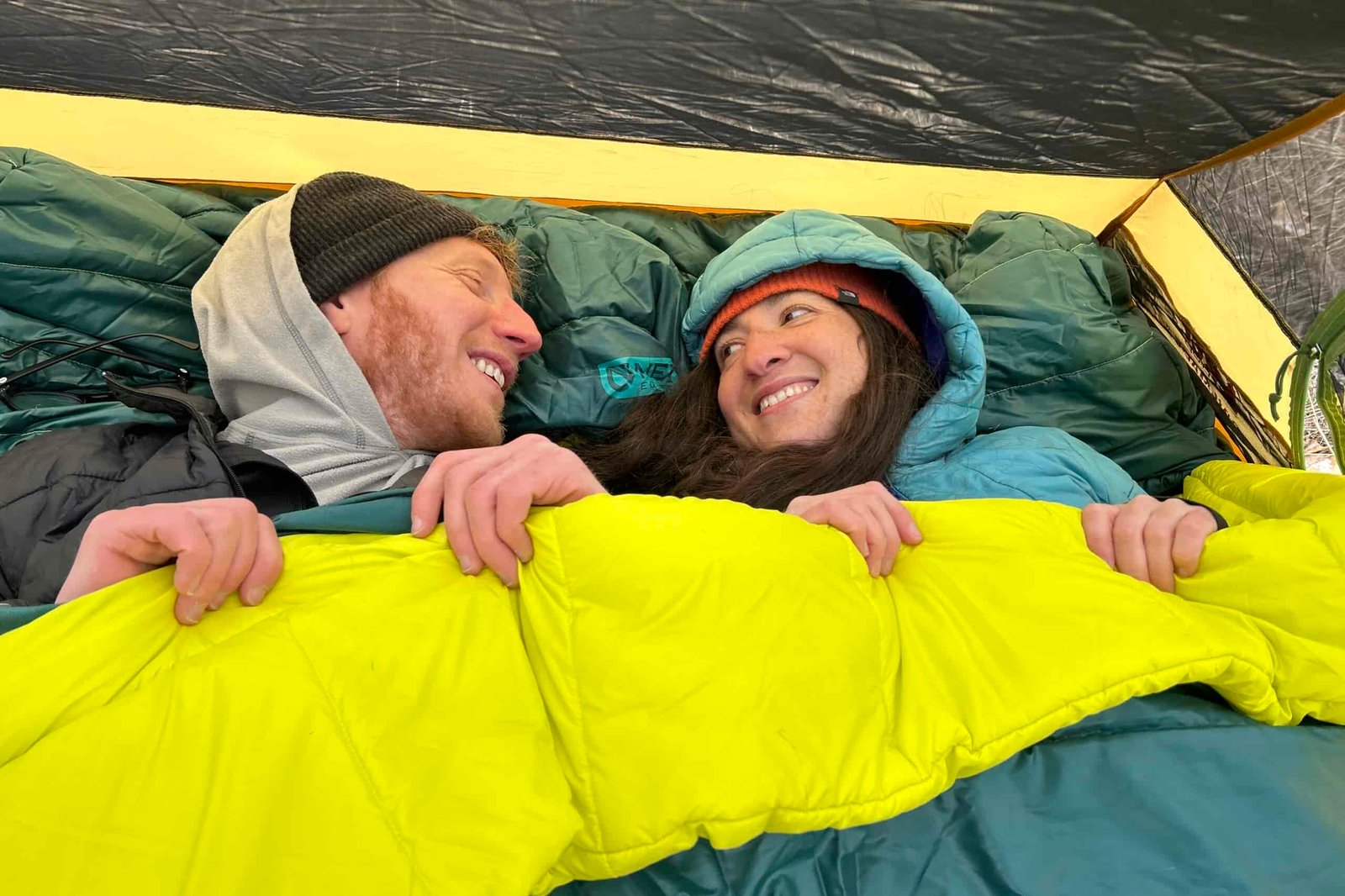 A man and woman smile at each other in a double sleeping bag in a tent.