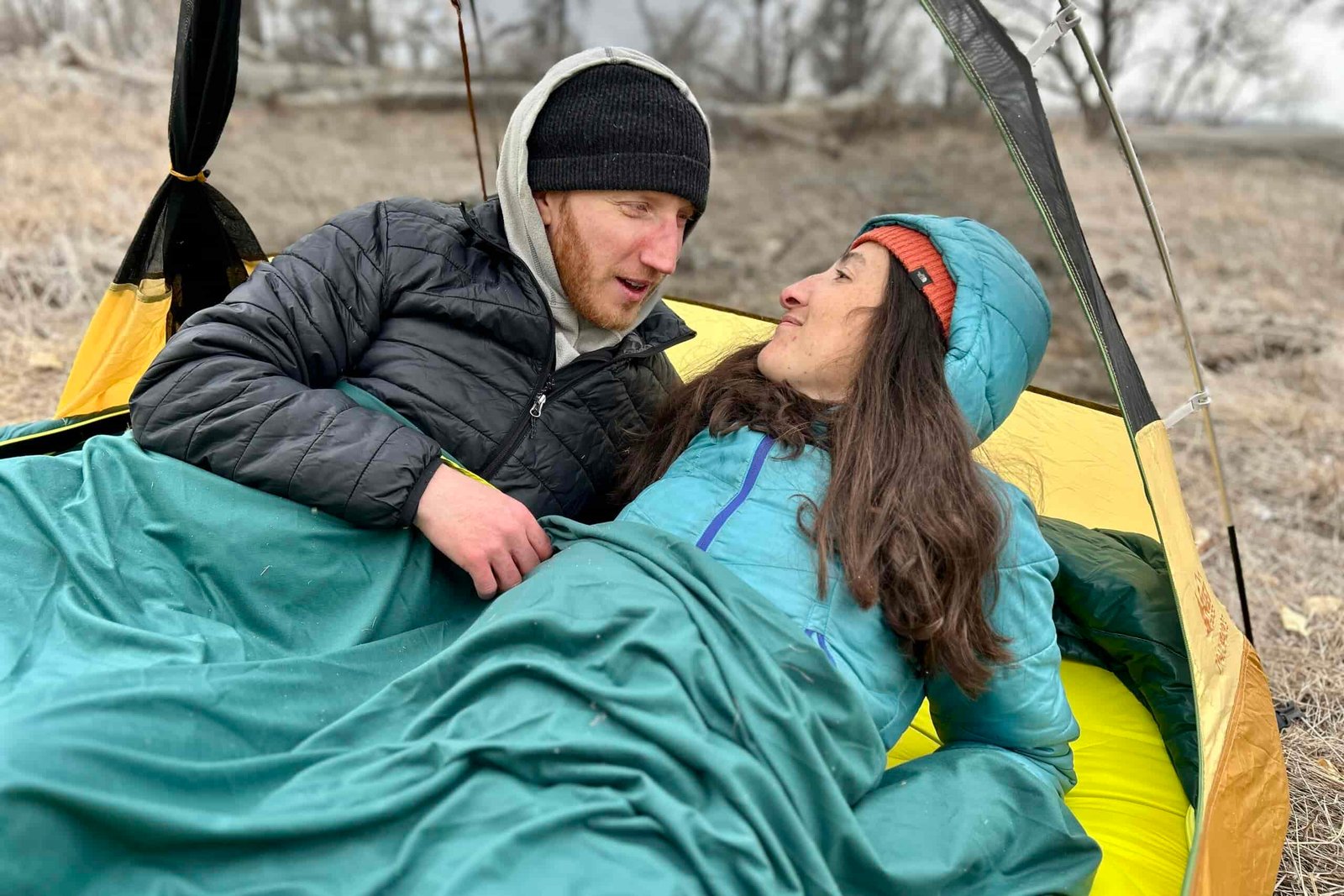 A man and a woman lay in a camping tent in a double sleeping bag talking.