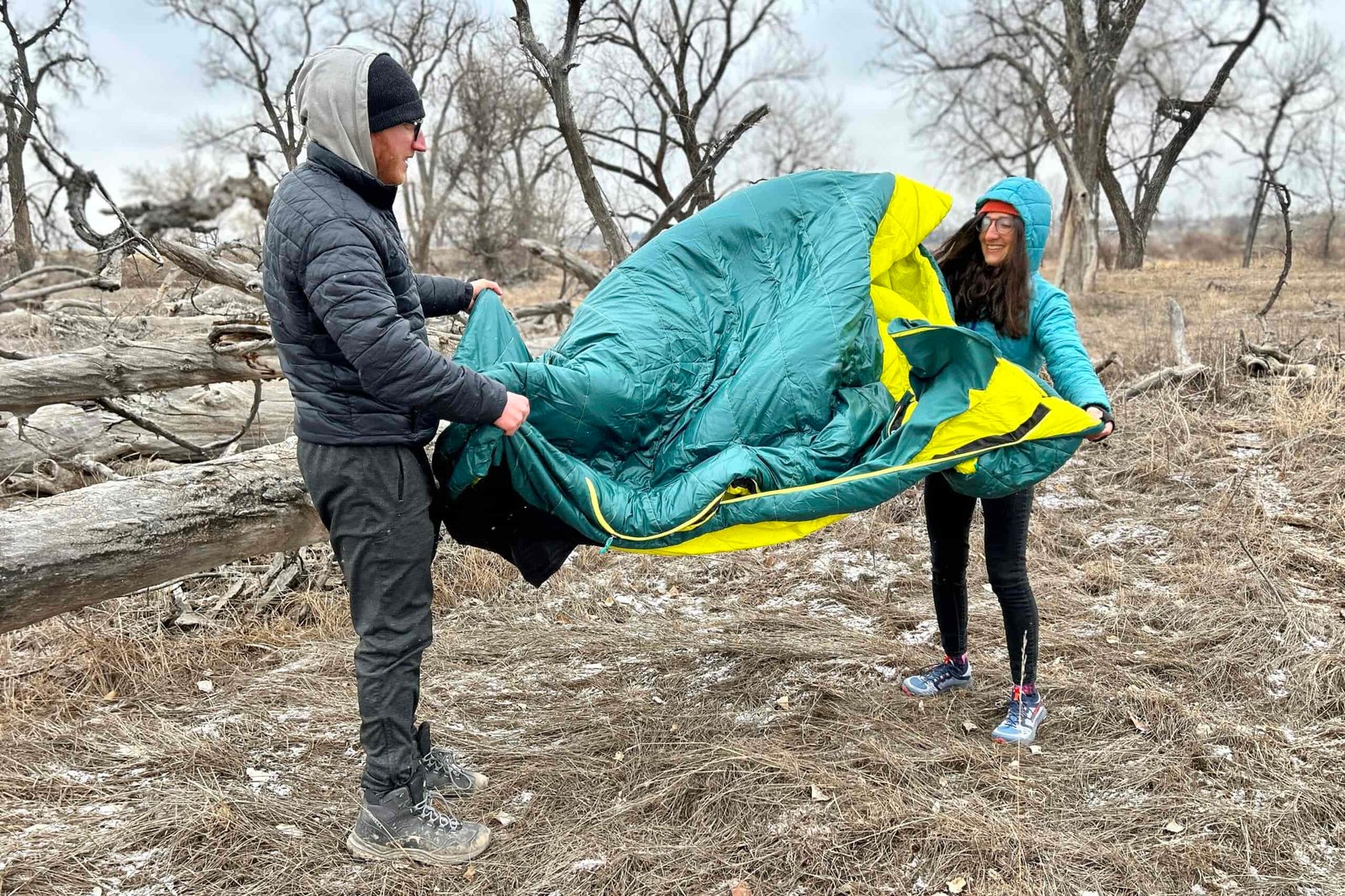 Two people fluff a double sleeping bag in a wintry forest setting.