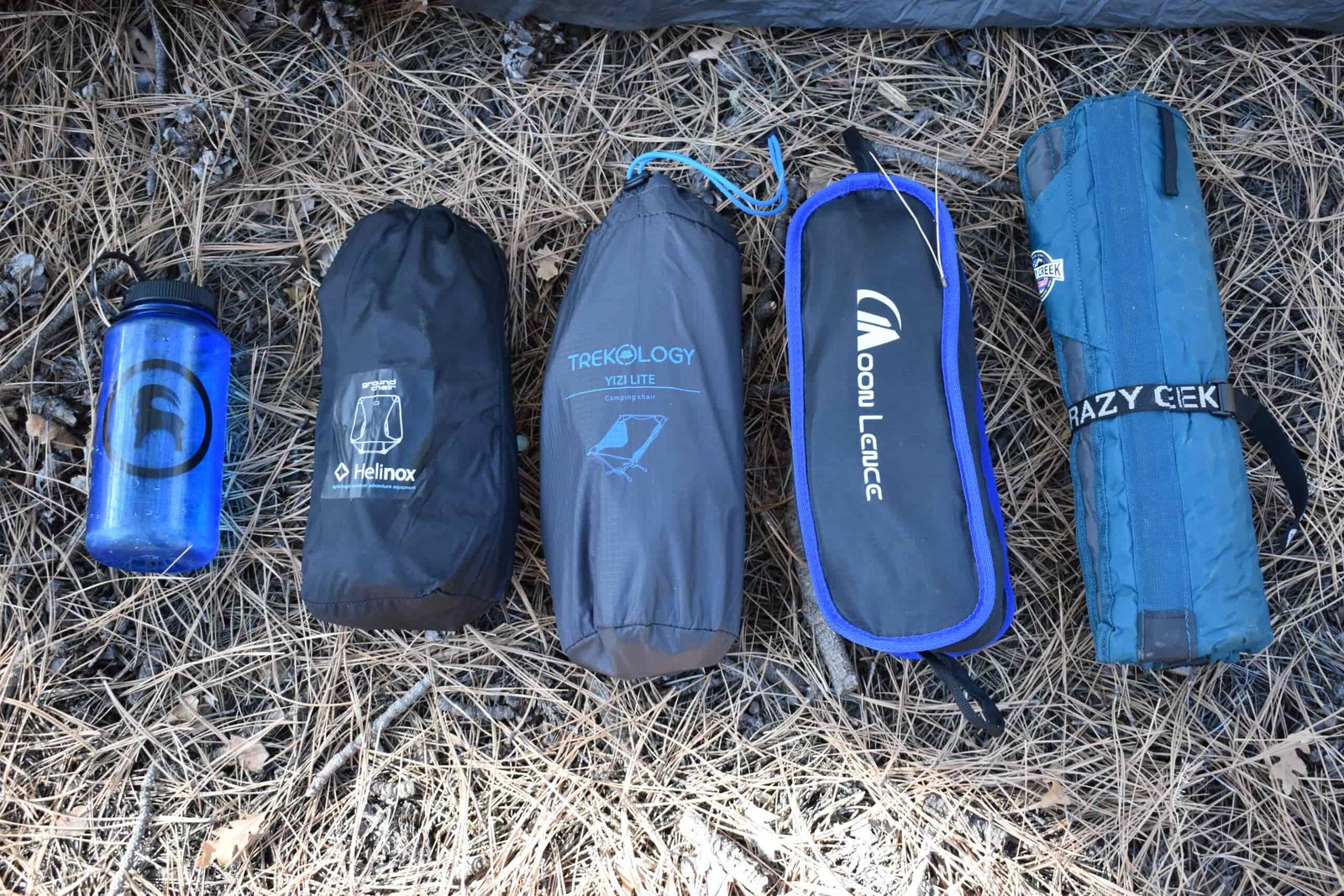 4 packed backpacking chairs lay next to a 1 L Nalgene bottle on a bed of pine needles to compare their packed sizes