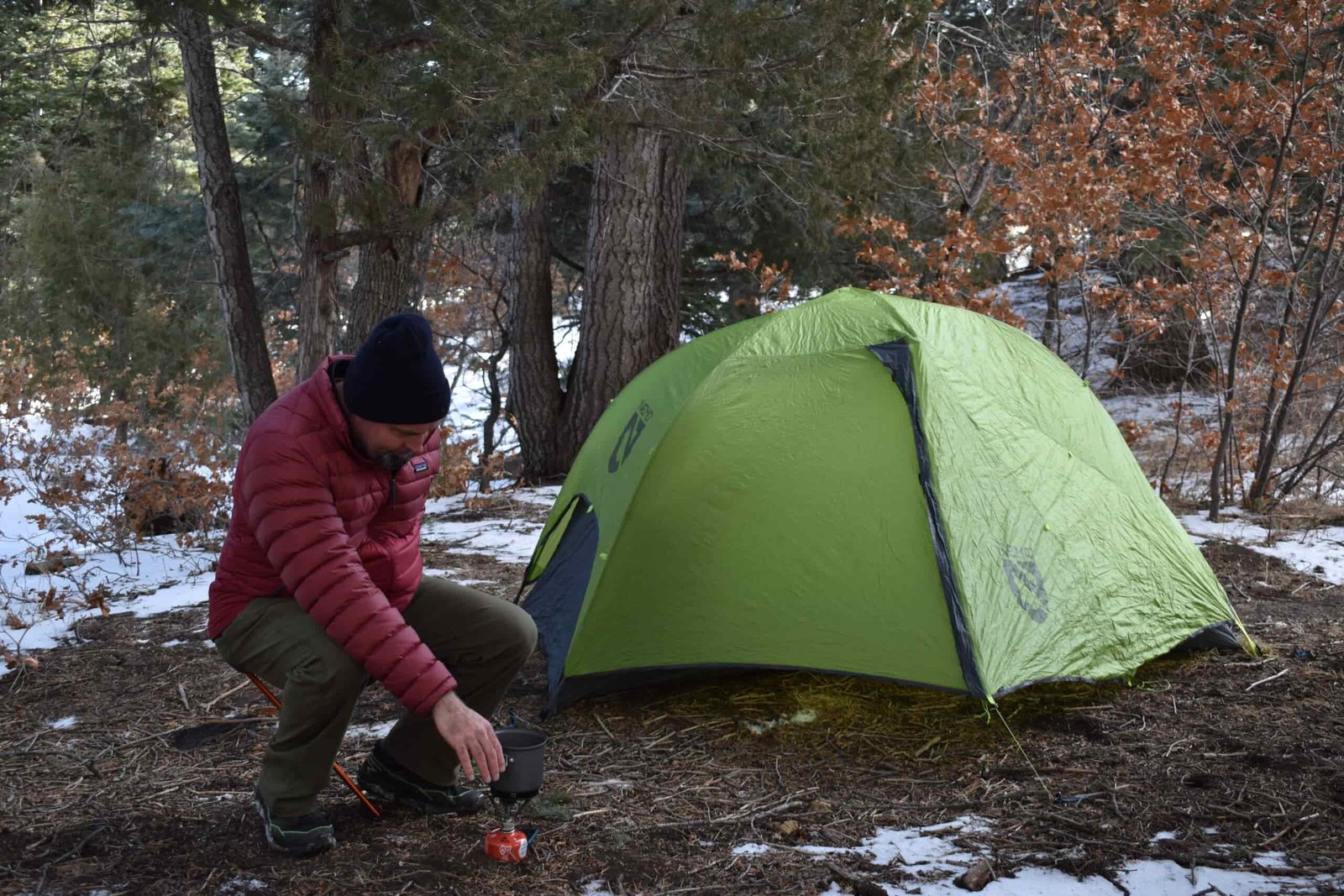 a camper sits on the stool as he leans forward to work with his camp stove outside of his tent