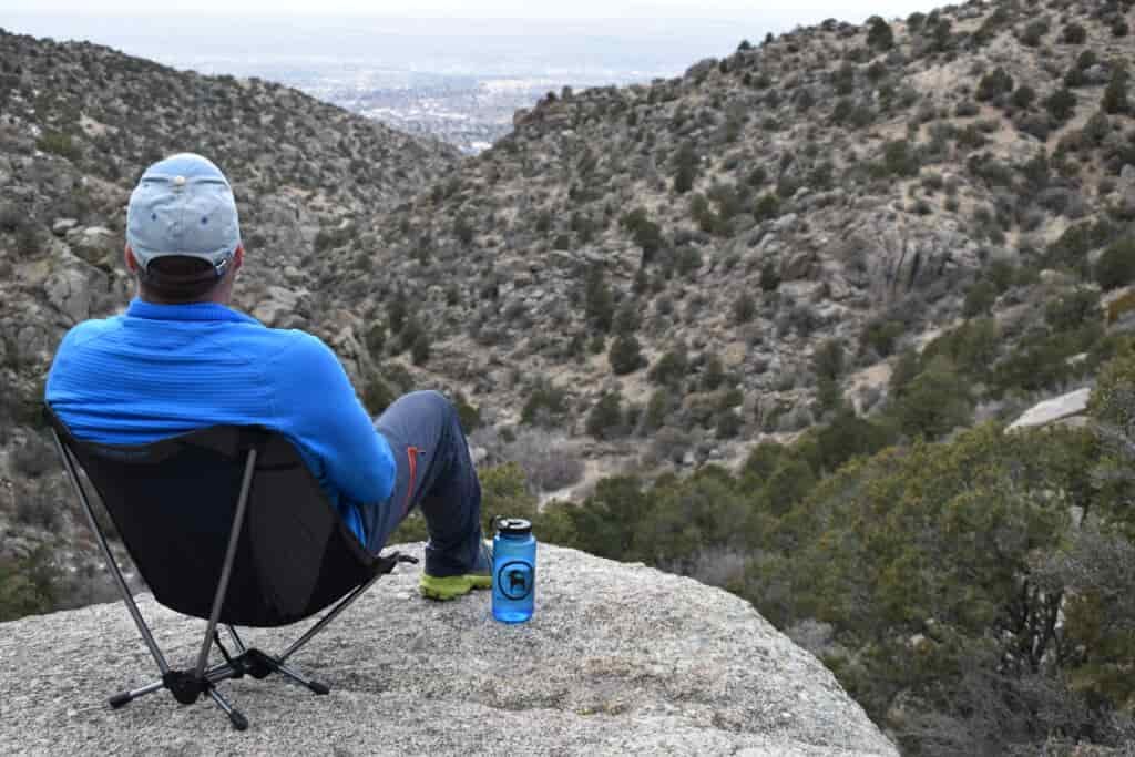 hiker sits on a rocky cliff overlooking a desert canyon while sitting on a camp chair
