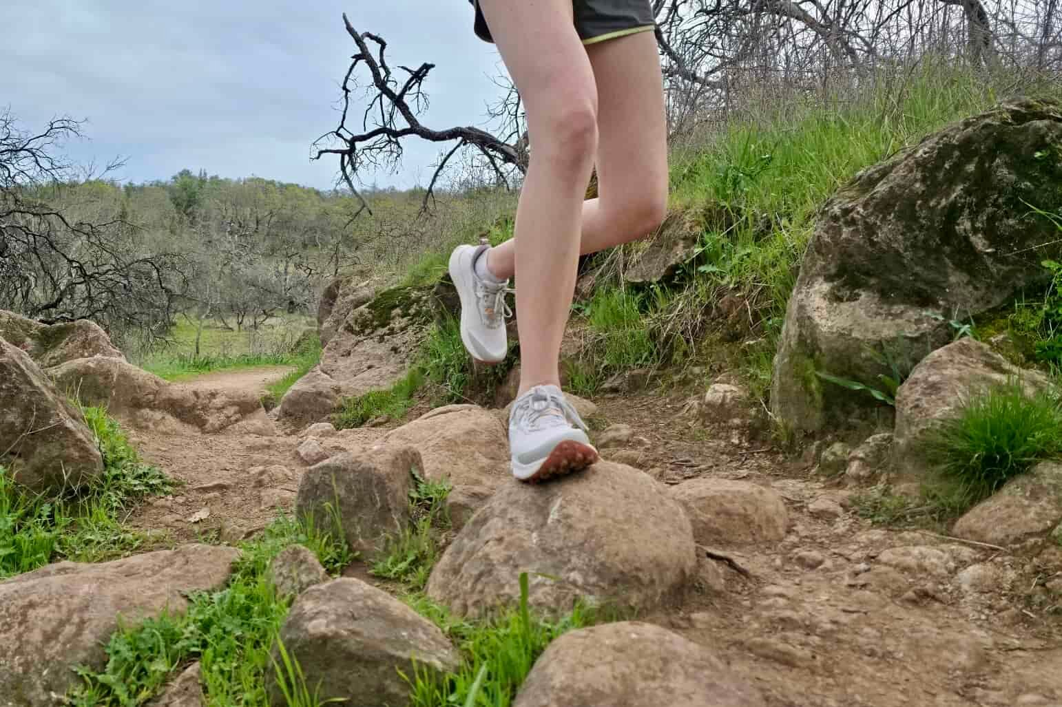 A trail runner runs along a rocky wooded trail in a pair of Saucony shoes.