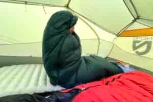 a hiker sits up in a tent, wrapped cozily in a sleeping bag