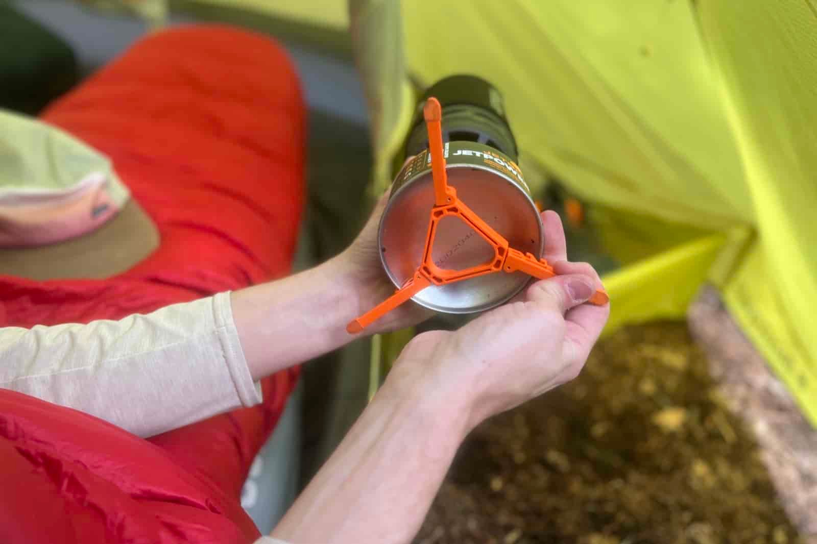 A close up of a hiker in a tent fitting a pot stabilizer to the bottom of a fuel canister.
