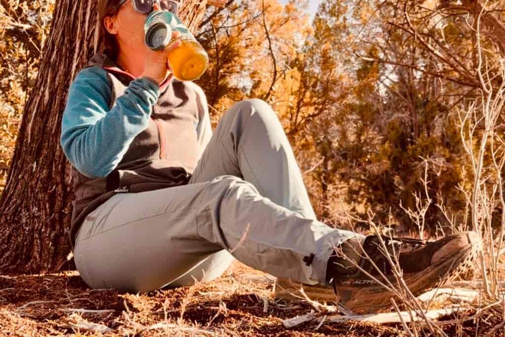A person wearing the Baleaf pants is resting against a tree in the woods drinking water from a water bottle.