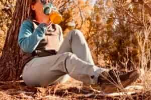 A person wearing the Baleaf pants is resting against a tree in the woods drinking water from a water bottle.