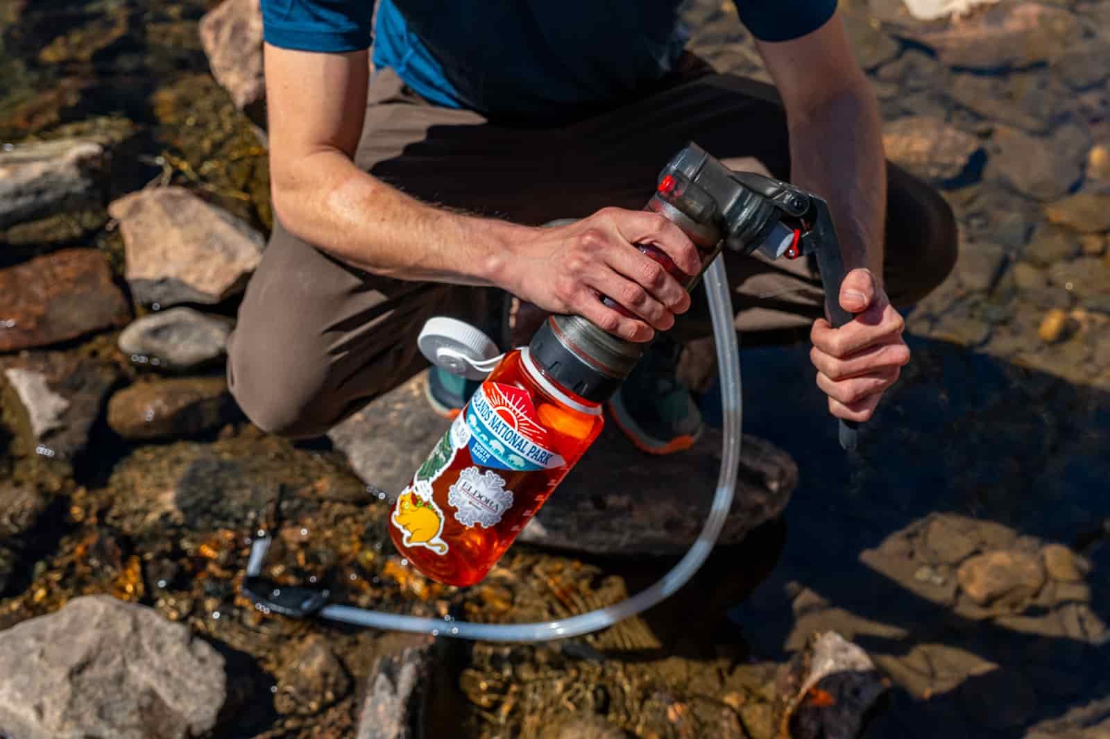 a hiker stoops on a rock in a creek while pumping water with the msr guardian and a nalgene bottle.