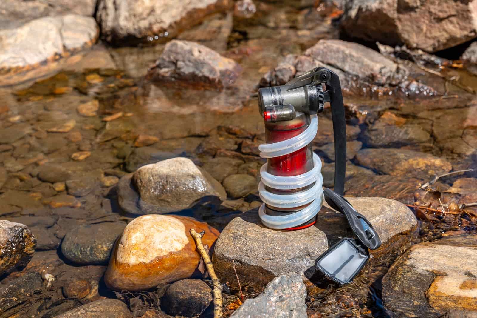 A pump water filter sits on a rock in a creek with the hose wrapped around it