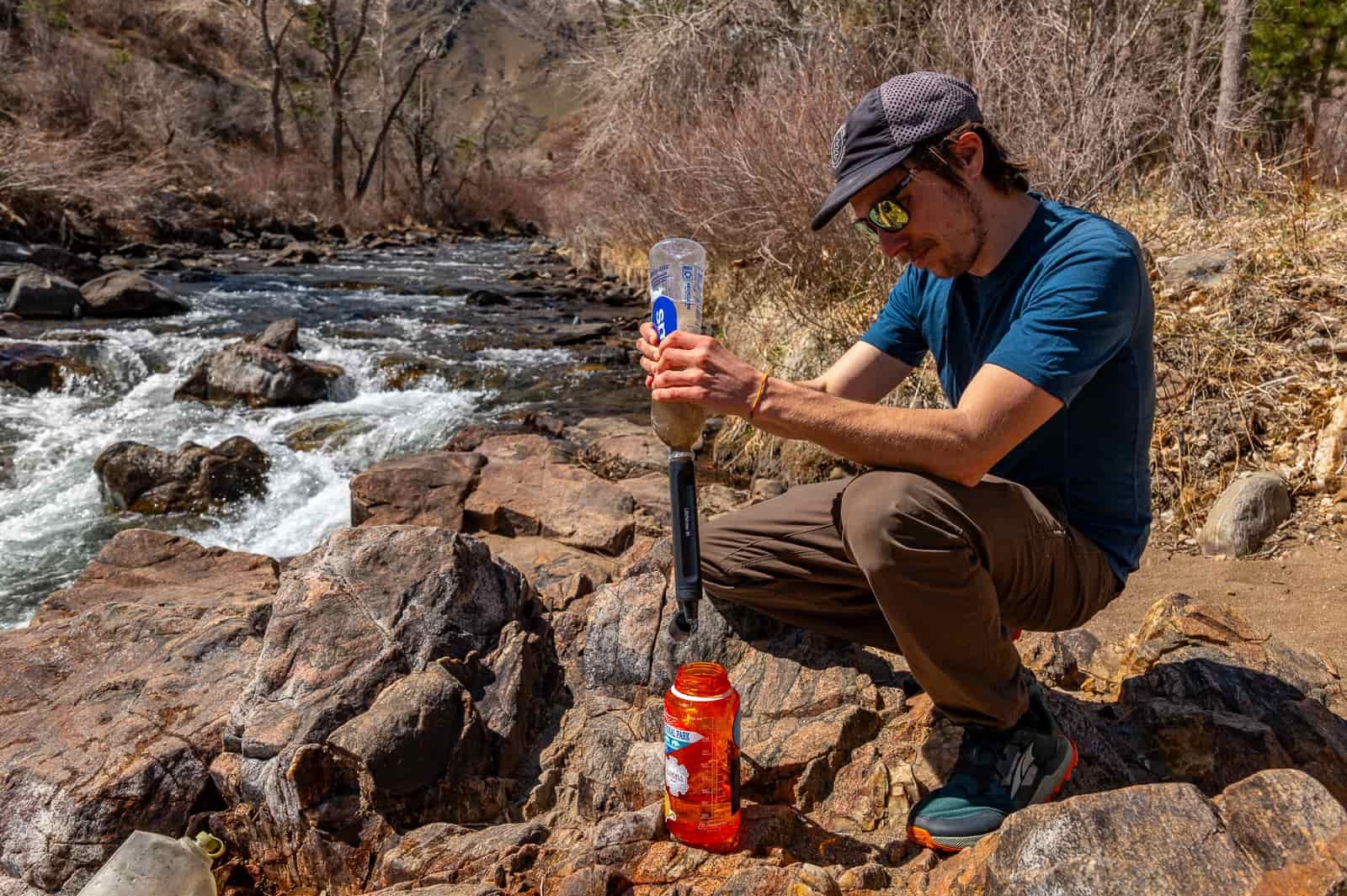 A hiker sits by a creek and squeezes water through the peak straw into a nalgene bottle.