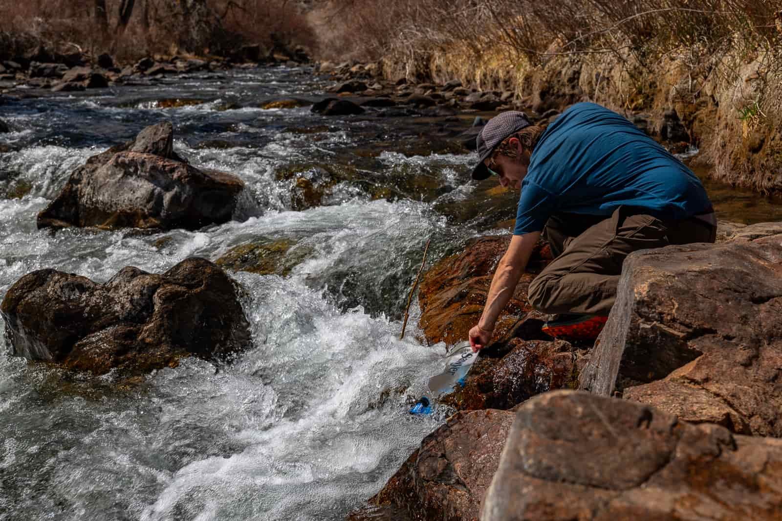 A hiker bends down to fill a bladder with creek water.