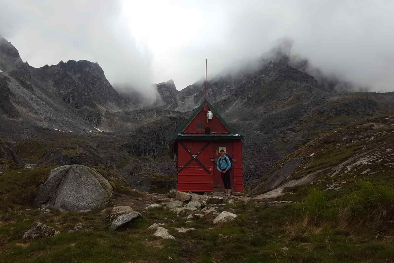 A woman stands in front of a red mountain hut.