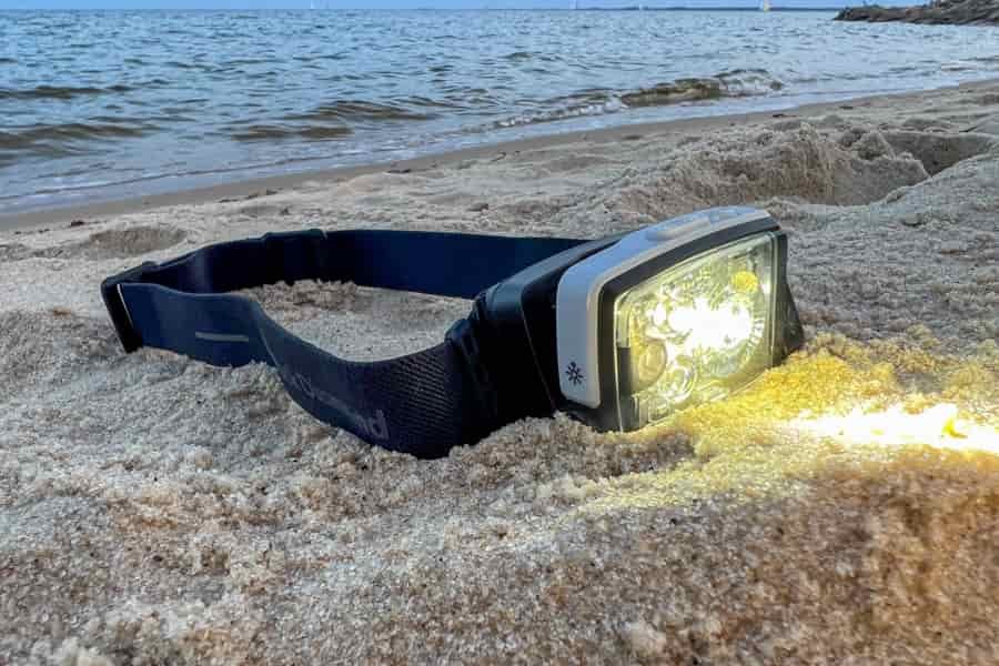 A headlamp sits in the sand near the ocean. The light is illuminated on the headlamp.