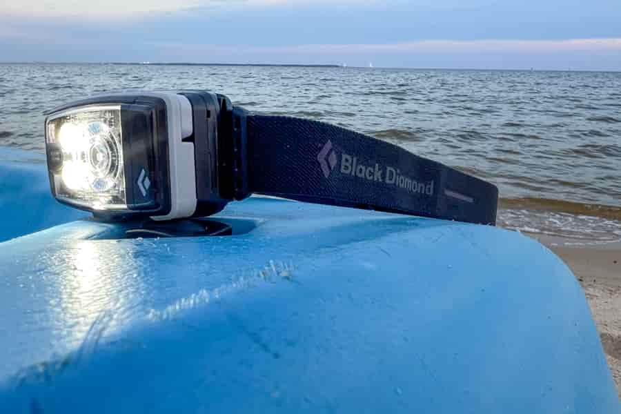 An illuminated headlamp sits on a kayak on the beach. The ocean is in the background.