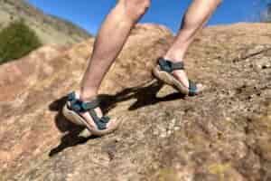 Close up image from knees down of a person ascending a steep sandy rock in hiking sandals