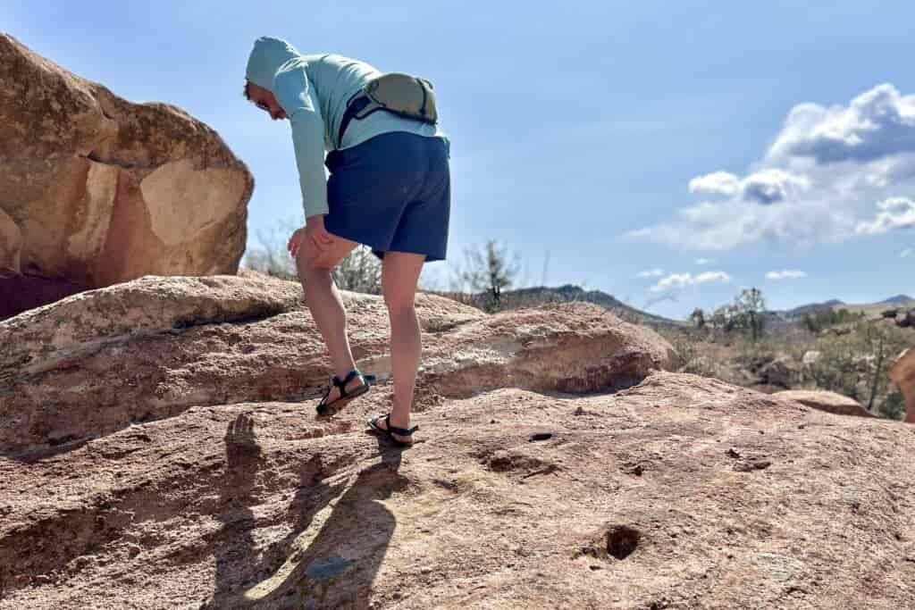 A man wearing a sun hoodie, shorts and sandals bends over as he climbs up a steep rock on a sunny day.