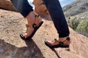 Close up image from knees down of a person in pants and sandals hiking up a steep rock on a sunny day