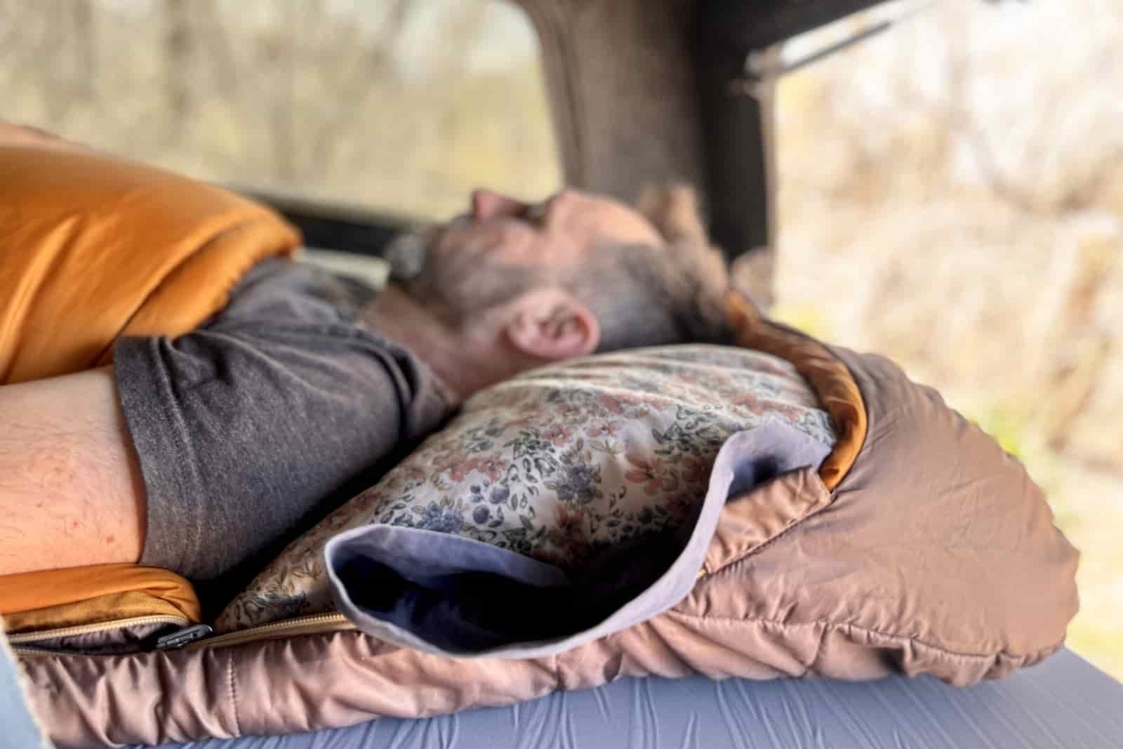 Profile image of a pillow tucked into the hood of a sleeping bag with a person laying on top, sleeping.