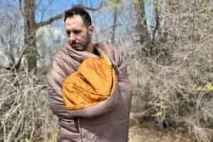 A man from the waist up is wrapped in a sleeping bag, standing outside and looking down at the ground