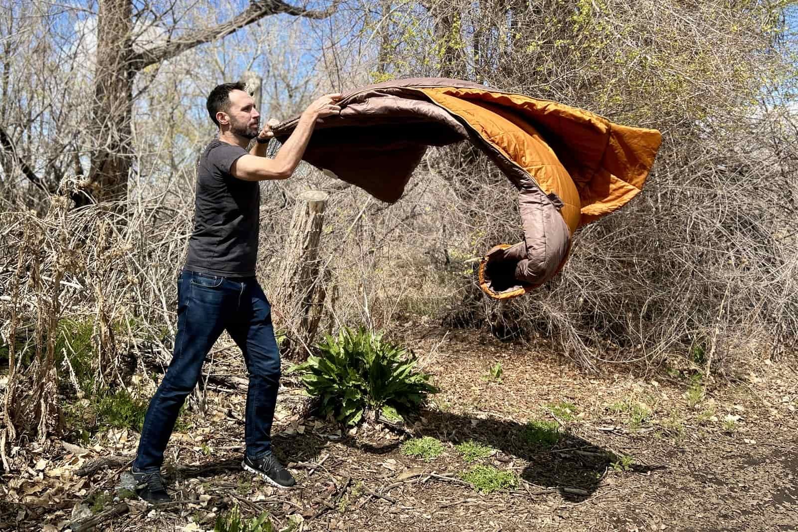 A man in a t-shirt and jeans fluffs out a sleeping bag in a forested setting.