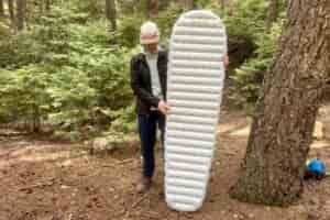 a hiker stands in the woods and holds up a gray sleeping pad.