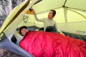one hiker lays in a tent while another reaches into an upper pocket above him