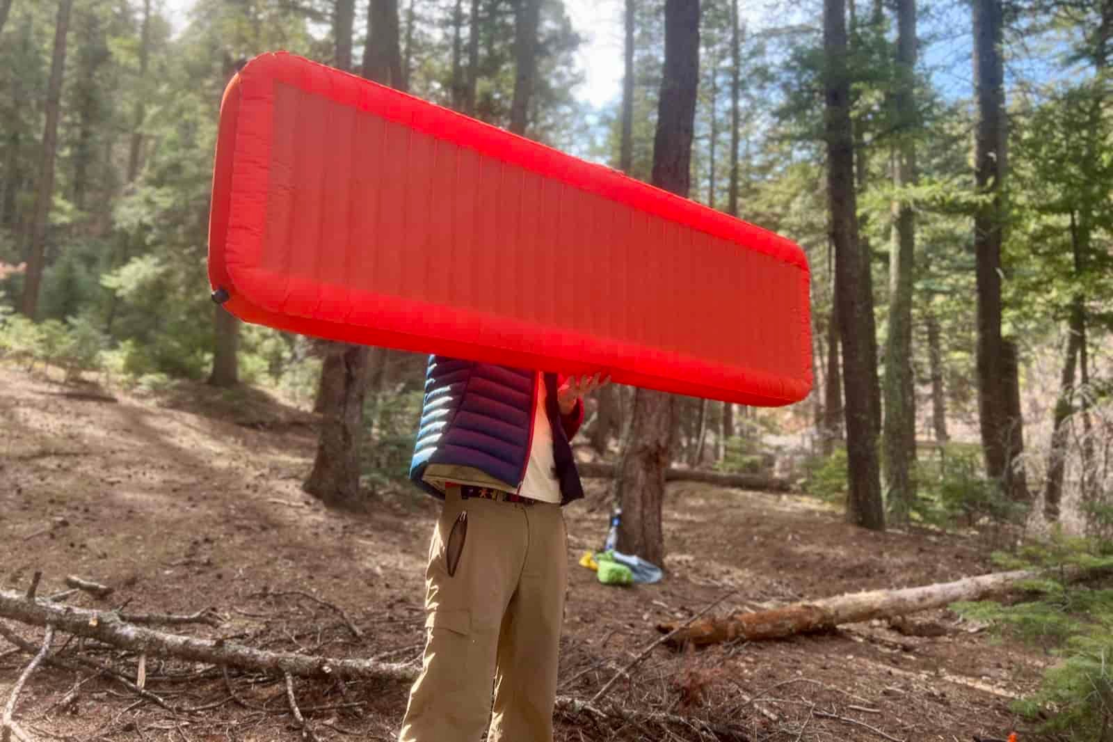 A hiker holds a red sleeping pad in the air in the middle of the woods