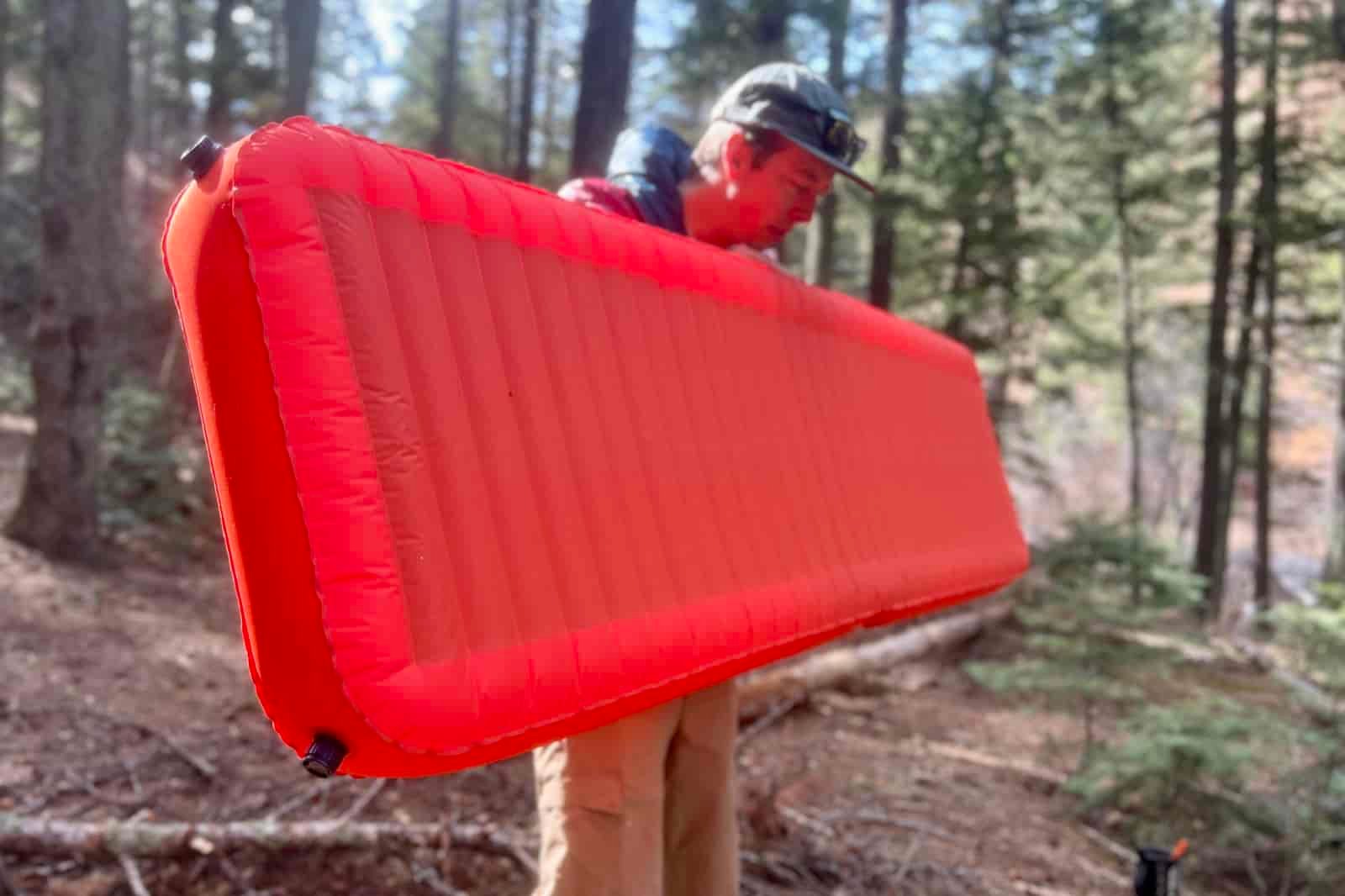 a hiker holds an inflated red sleeping pad in the middle of the woods