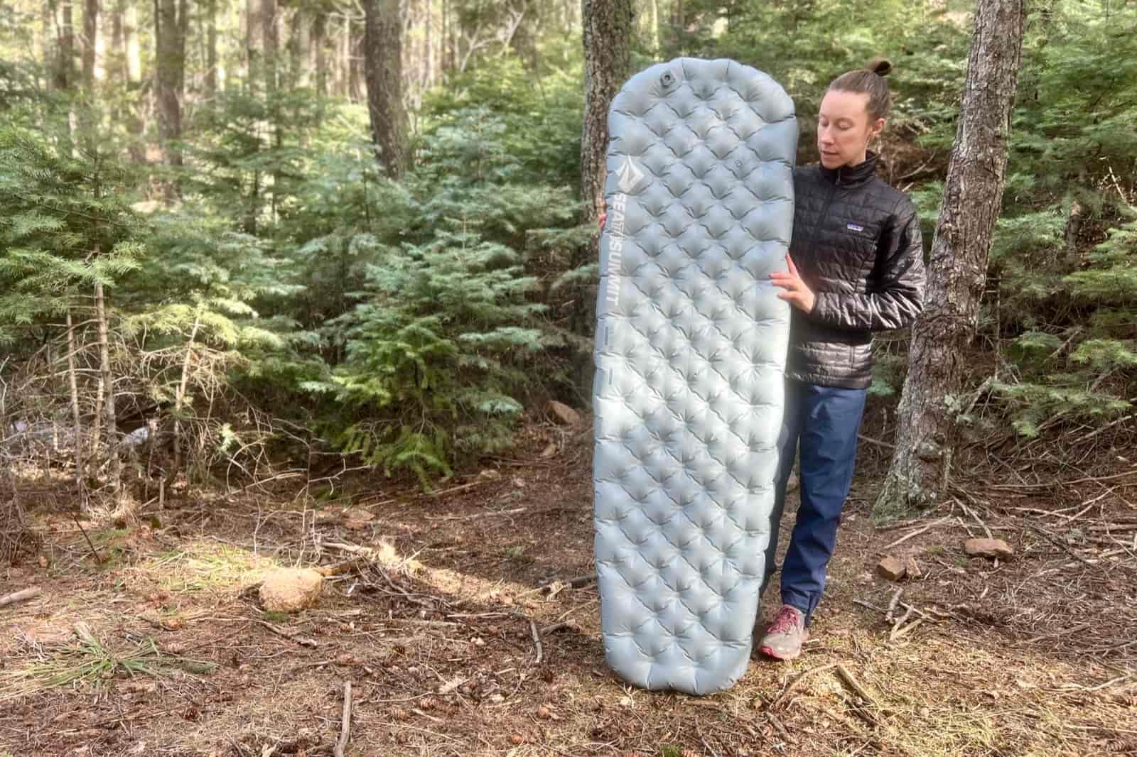 a hiker stands in the woods and holds up a gray sleeping pad.