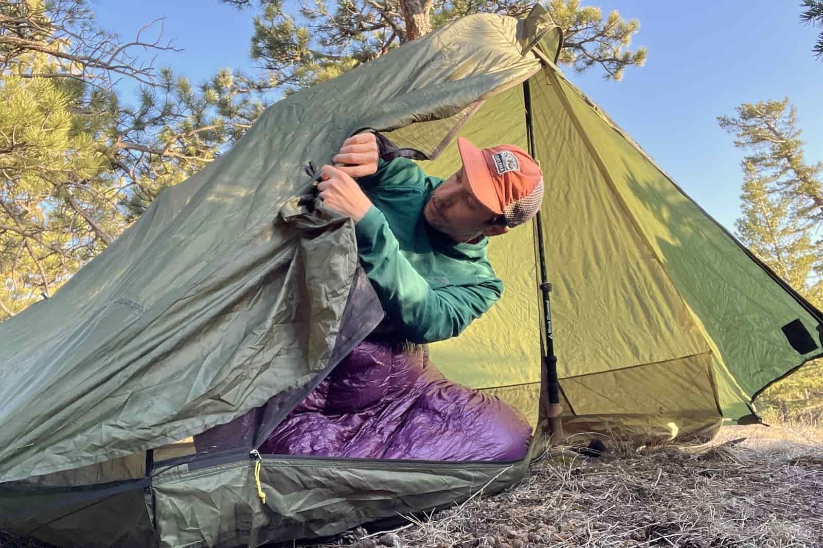 a hiker leans out of a green trekking pole tent to tie back the door